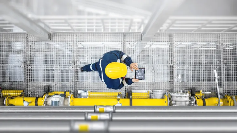 Engineer wearing a hard hat reviewing a tablet beside labelled industrial pipework on a guarded access platform